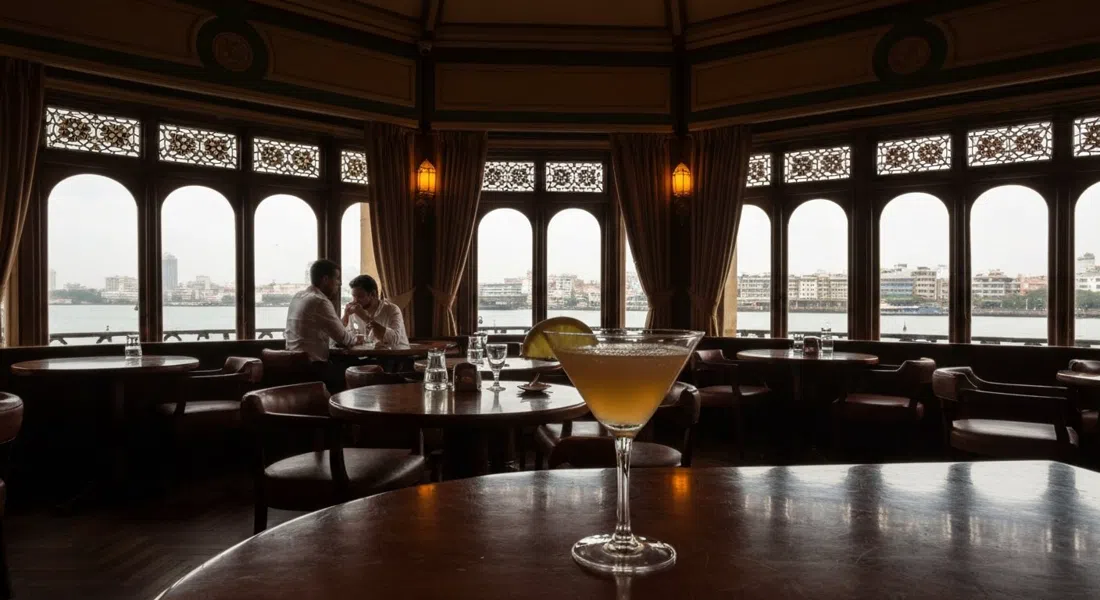 el interior Art Decó tenuemente iluminado de Harbour Bar dentro del Taj Mahal Palace, una copa de cóctel en primer plano y vistas al puerto a través de las ventanas